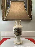 Full view of vintage porcelain table lamp with beige lampshade and ornate painting on the base.