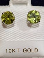 Close-up photo of both round faceted light green peridot earrings set in 10K gold prong settings displayed on a white textured holder with '10K T. GOLD' printed beneath.