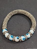 Flexible metallic mesh bracelet with alternating white beads featuring blue and black eye designs combined with rhinestone spacer rings.