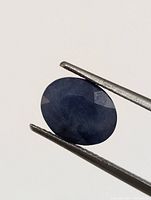 Close-up image of one oval-shaped sapphire held by tweezers showing dark blue color and faceting.