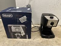 DeLonghi Stilosa espresso machine beside its original box. Black and silver design with control knob on front and steam wand left side.