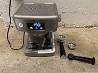 Front view of the espresso coffee machine showing digital display at 198°F, power button lit, steam wand, drip tray, portafilter handle, filter basket, and tamper laid next to the machine.