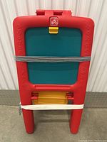 Red plastic Step2 kids easel folded and strapped closed, showing green chalkboard side with black and white cord securing it.