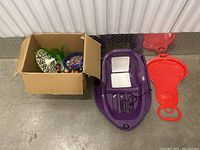 Photo of two sleds (purple and red) next to a box filled with various small toys on a concrete floor.