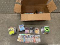 Large cardboard box with multiple stacks of Pokémon cards including energy cards, loose and sleeved, spread out in front of the box.