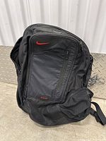 Front view of black Nike backpack with red swoosh logo, multiple zippered compartments visible including front diagonal zipper and side pocket.