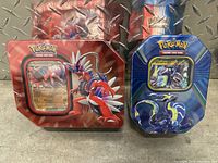 Front view of two sealed Pokemon tins on metal textured surface, featuring Koraidon on the red tin and Miraidon on the blue tin.