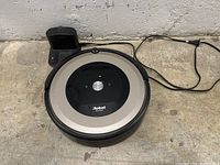 iRobot Roomba robot vacuum cleaner with charging dock and power cable sitting on concrete floor.