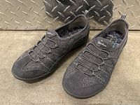 Pair of black/dark gray Skechers women’s shoes on a metal surface, showing upper side and elastic lace design.