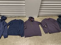 Four sweaters laid on the floor against a wall, showing various pullover and crewneck styles in navy and purple colors.