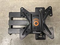 Close-up image showing a black metal TV wall mount bracket with a central X-shaped plate and three extended arms. The mount has multiple holes for attaching to a wall and TV, showing an orange circular logo and caution text.