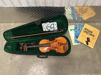 Full view of violin inside an open hard case with case lining in green, bow secured above violin, violin shoulder rest in adjacent compartment and three violin instructional books outside the case.