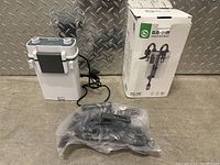 Photo showing white and gray external aquarium water filter unit with black power cord and various accessories sealed in a plastic bag next to the unit.