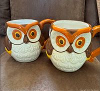 Image of two ceramic coffee cups shaped like owls with orange, brown, white, and yellow painting, sitting on a brown surface.