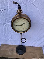 Front view of the antique-style brass clock on a black metal stand with a round base and valve handle design.