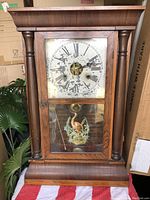 Front view of antique wooden mantle clock showing worn clock face with Roman numerals and painted bird image on lower glass panel, framed by columns and topped with crown molding.