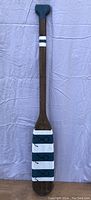 Full view of the wooden boat oar painted with white stripes and fitted with three black hooks on the blade end, laid on a light fabric surface.