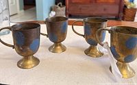 Four brass cups displayed on a white surface with a brownish patina and tarnish visible, each cup having a handle and pedestal base.