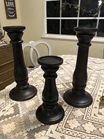 Three black wooden candlestick holders of varying height displayed on a patterned tablecloth with a window and chairs in the background.