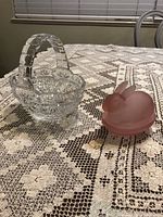 Both items on a crocheted lace tablecloth: clear crystal basket cake plate to the left and pink rabbit candy dish to the right, closed.
