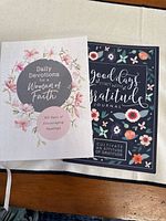 Two devotional journals placed on a wooden surface. The left one titled 'Daily Devotions for a Woman of Faith' features a light-colored floral cover with pink flowers. The right one titled 'Good Days Start with Gratitude Journal' has a dark blue floral cover with pink and orange flowers.