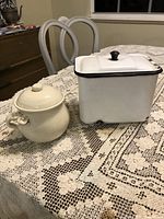 Photo of enamelware refrigerator box and ceramic jar on lace tablecloth, showing front and side views with lids on.