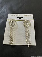 Photo of a pair of dressy formal fashion jewelry earrings on a white card labeled 'FASHION JEWELRY'. The earrings have circular rhinestone studs and three hanging chains of rhinestones each.