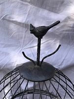 Close-up of the bird-shaped finial atop the cage showing detailed metalwork and branch-like hooks