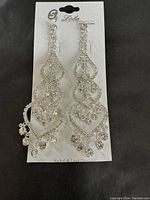 Photo shows a pair of Lola brand silver-tone chandelier earrings with multiple rhinestone loops and dangling rhinestone drops attached to branded card.