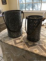 Two metal luminary lanterns with heart-shaped cutouts and rope handles sitting on lace tablecloth with window in background.