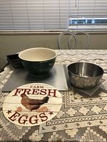 Photo showing the 10.75 inch mixing bowl, stainless steel mixing cup, rectangular metal baking sheet, small dark metal loaf baking pan, and 'Farm Fresh Eggs' round wooden sign on a table with lace tablecloth under a window with blinds.