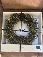 Olive leaf wreath placed inside a white box with black ribbon tied across it. The box has a label showing the brand Pimpernel and the product name Olive Wreath.