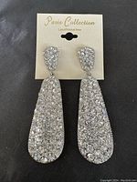 Photo shows a pair of long teardrop shaped dangle earrings fully covered in clear rhinestones on a silver-tone metal base, attached to a card labeled 'Paris Collection'.