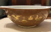 Close-up side view of large Pyrex mixing bowl showing brown exterior and golden-yellow Early American pattern of birds and kitchen motifs.