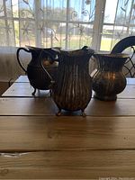 Three silver-plated water pitchers arranged on a wooden surface in front of a window with sheer curtains.