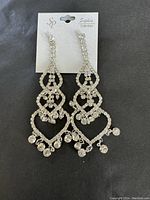 Photo of a pair of silver-tone dressy formal earrings with clear rhinestones in heart-like and swirling design, hanging on a Sophie Collection branded card.