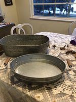 Both metal items on a lace tablecloth, showing oval metal tray with wooden handles and oval wash tub behind it.