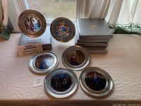 Six Jefferson pewter plates displayed on table, some upright and others laying flat, with original grey boxes stacked to the side.