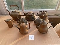Photo showing all eight copper teapots arranged on a table, various sizes and handle styles visible.