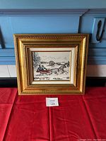 Frontal view of ornate gold-framed winter sleigh print resting against blue paneling and red cloth.