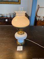 Electric hobnail milk glass hurricane lamp with brass accents and glass chimney shown illuminated on a table with other white dishware and furniture in background, photo taken for lot detailing.