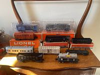 Overview of ten Lionel O-gauge train pieces displayed on table: two sealed cars, orange boxed car, loose freight cars, passenger car, tank car, steam locomotive and tender.
