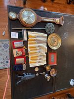 Overview of all lot items on black wooden table including barometer, flatware, decorative plate, U.S. star grave marker, silver spoons, insect figurine, and match safes.