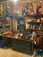 Full view of the workbench with various tools, tool cases, and a black multi-drawer tool chest underneath. Multiple cords hanging overhead.