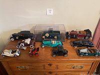 Wide view of all 10 model cars arranged on top of a wooden dresser, showing variety of colors and styles.