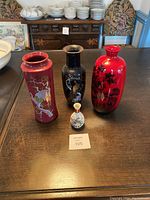Four vases displayed on a wooden table: three tall vases in maroon, black, and bright red colors with various floral and bird designs, and one small white Satsuma vase with detailed painting.