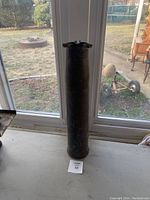 Full view of the tall copper military shell casing standing upright on a table near a window, showing the overall shape and patina.