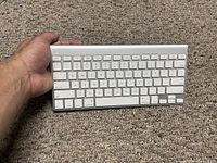 Apple Bluetooth keyboard Model A1314 held in hand showing front key layout, on carpet background.