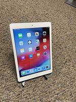 Apple iPad Air displayed on a carpet floor showing apps on screen, held vertically on a black stand.