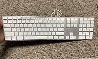 Top view of Apple USB wired keyboard on carpet background showing full key layout and attached USB cable.